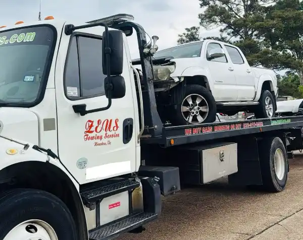 Zeus Cash For Junk Cars tow truck hauling a wrecked white pickup truck in a Cypress, TX neighborhood.